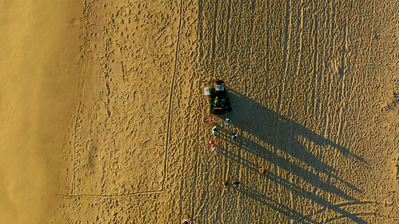 Overhead Diamante Cabo San Lucas beach frame showing the cart and people against dense sand texture, with straight track lines and elongated shadows crossing the image.