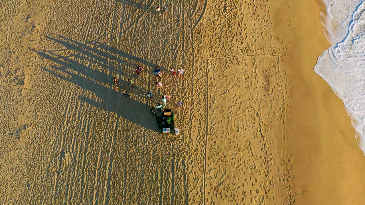 Overhead drone frame in Diamante Cabo San Lucas showing a small group on textured sand, long shadows stretching left, and white ocean foam along the right shoreline.