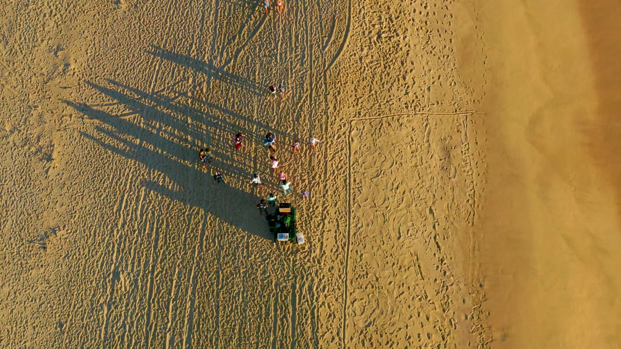 Top-down aerial frame in Diamante Cabo San Lucas showing a small group of people beside a compact vehicle on textured sand, with long shadows extending across the beach.