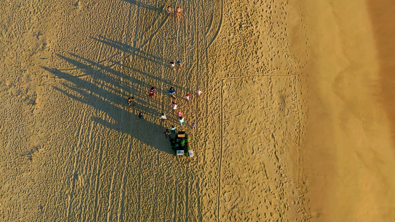 Top-down view in Diamante Cabo San Lucas showing the beach group and vehicle slightly right of center, with textured sand, faint tracks, and long shadows dominating the left side.