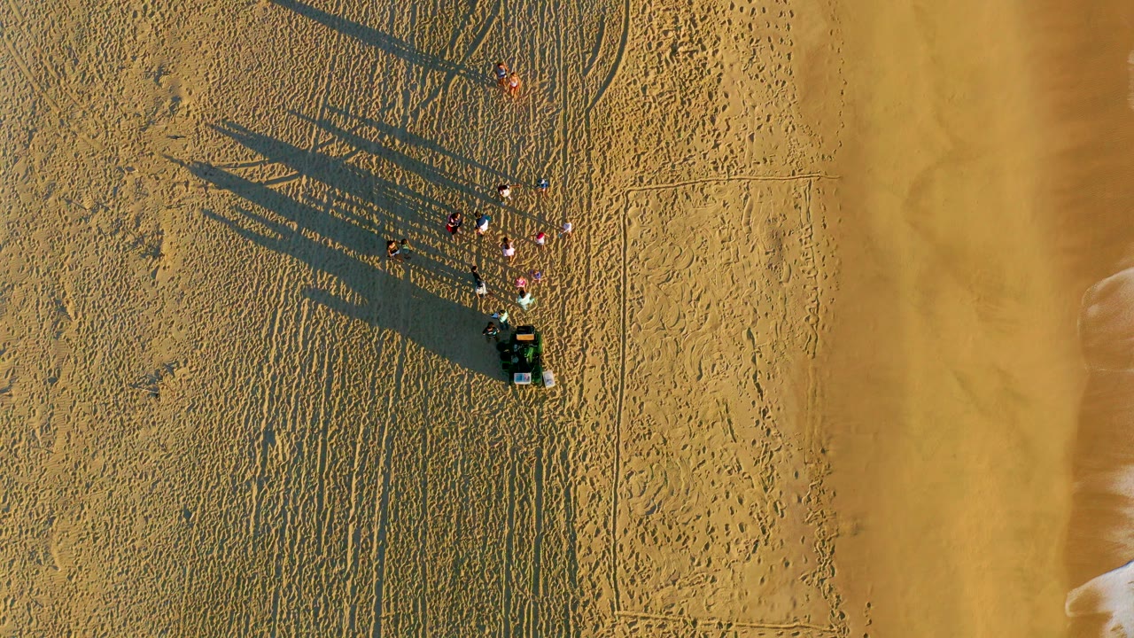 Overhead frame from Diamante Cabo San Lucas showing a small group of people and a dark vehicle on textured sand, with long shadows reaching left and smoother wet shoreline sand to the right.