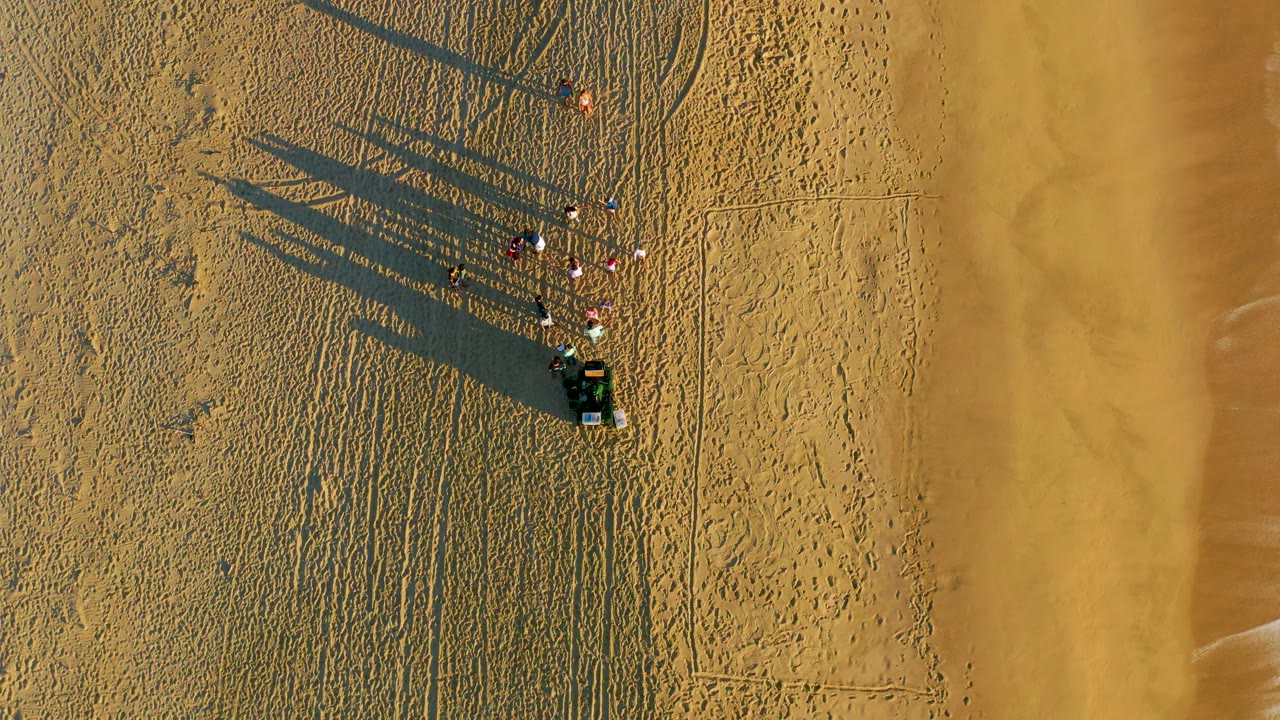 Top-down Diamante Cabo San Lucas beach frame with the people and vehicle still near the upper sand area, their shadows crossing textured sand toward a broad strip of smooth wet beach on the right.
