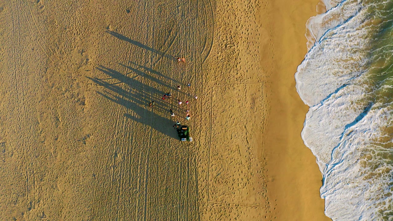 Overhead view at Diamante Cabo San Lucas showing a small group of visible people beside a dark cart on golden sand, with long shadows extending left and ocean foam along the right edge.