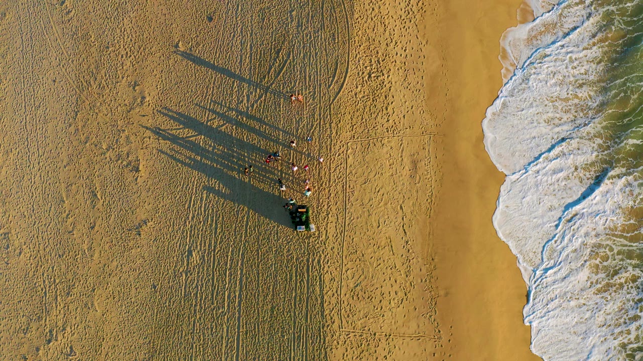 Aerial sand view with a group and cart casting long shadows near the surf.