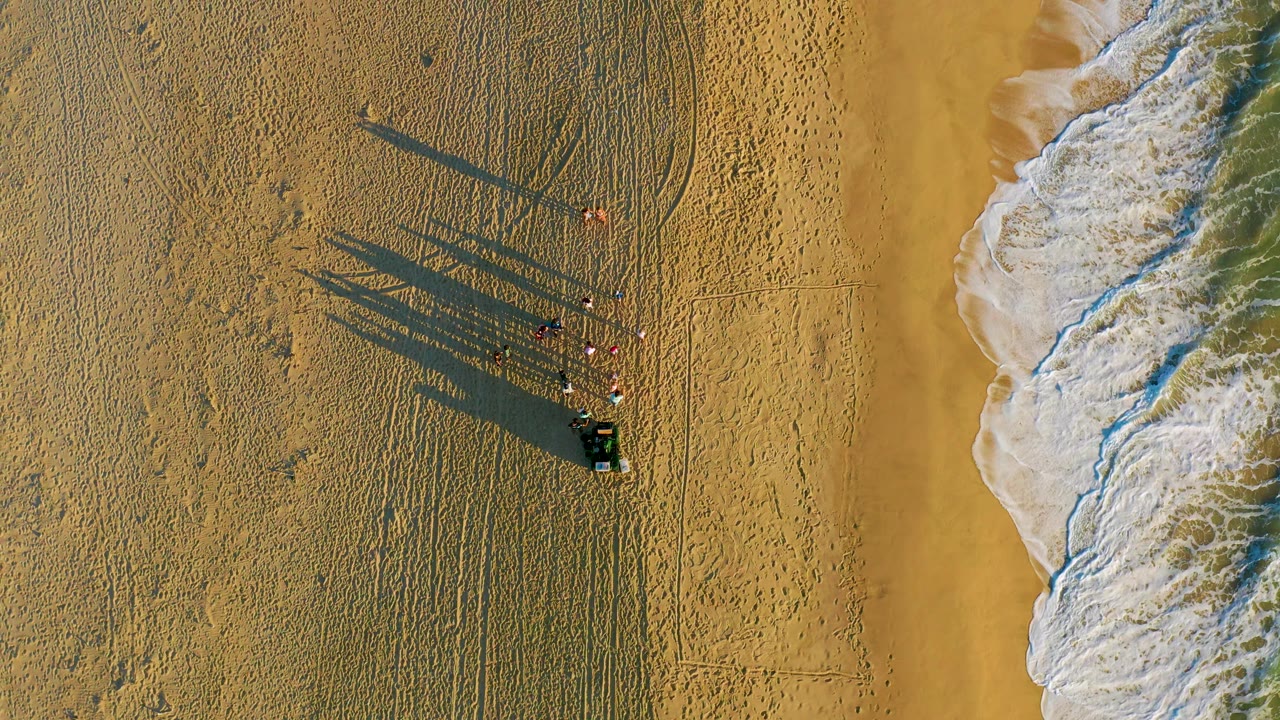 Top-down Diamante Cabo San Lucas beach view with visible people and a dark cart on patterned sand, while a broader band of white surf rolls down the right side of the frame.