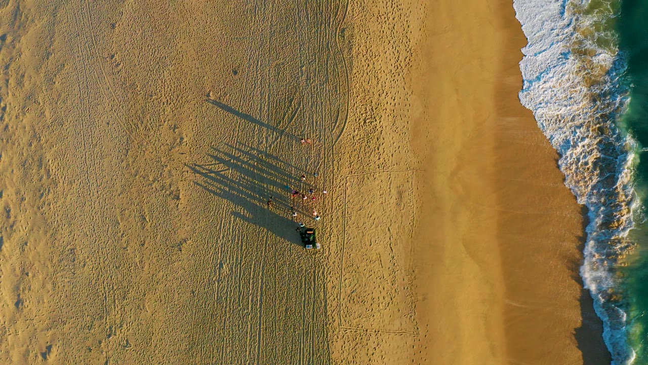 Overhead frame from Diamante Cabo San Lucas shows beachgoers standing on rippled sand near a dark object, with long shadows stretching left and white surf entering from the right.