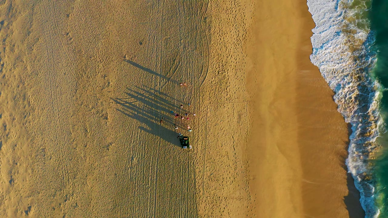 At 0:01, the aerial view holds the group near the lower left of the surf line as white foam thickens along the right side of the Diamante Cabo San Lucas beach.