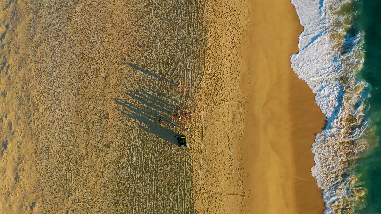 At 0:02, the people remain clustered on the sand while the wave edge curves more strongly into the right side of the overhead beach frame.