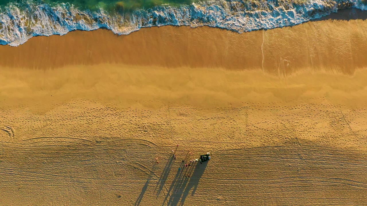 Overhead view of waves reaching the upper edge of a golden beach in Diamante Cabo San Lucas, with a small vehicle and long shadows on dry sand below, no visible people.