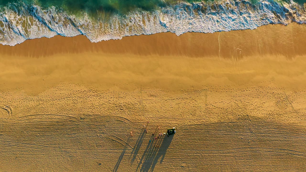 Foamy waves spread across the upper third of the Diamante Cabo San Lucas shoreline, with raked sand patterns, vehicle tracks, and a small vehicle casting a long shadow below, no visible people.