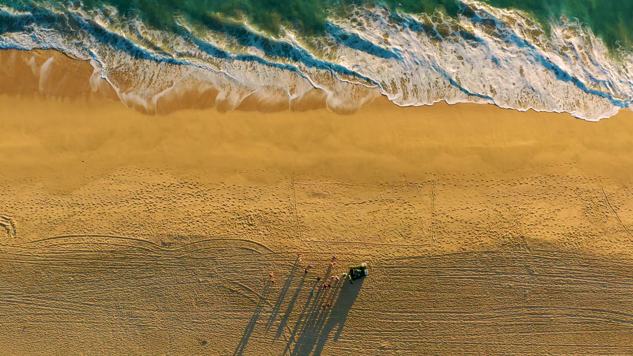 Overhead view at Diamante Cabo San Lucas showing foamy ocean at the top, broad sand below, a small dark vehicle, tiny people, tire tracks, and long shadows near the lower beach.