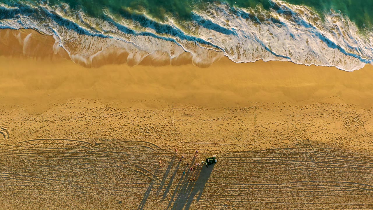 Drone view of waves, sand, vehicle, people, and long shadows.