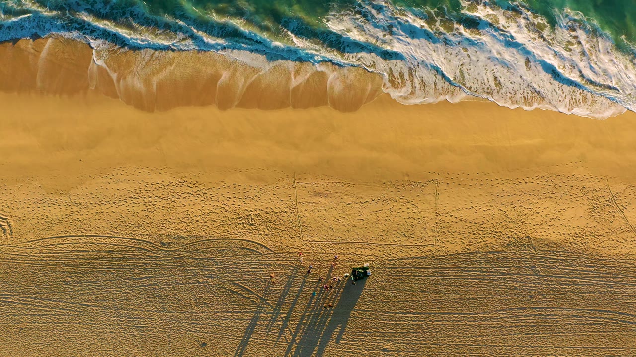 At 0:02, the overhead frame shows the surf pressed near the upper edge, open yellow sand through the center, and a dark vehicle beside tiny people and elongated shadows near the bottom.