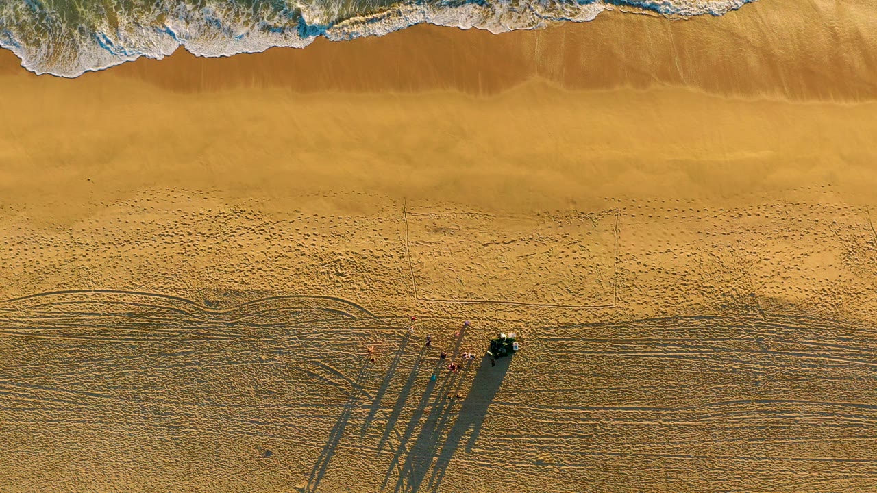 Overhead frame from Diamante Cabo San Lucas shows white surf at the top, wide golden sand below, tire tracks, a small group of people, a dark vehicle, and long shadows stretching across the beach.