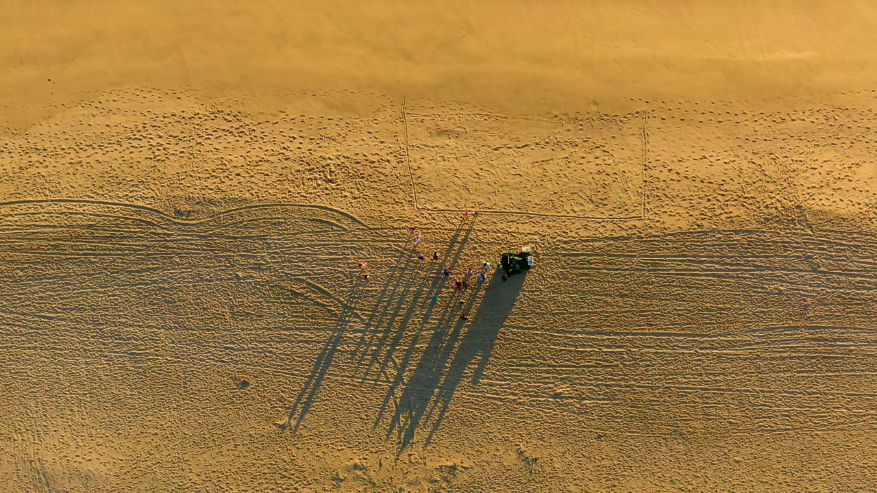 Overhead view of sandy beach in Diamante Cabo San Lucas with a small group of people, a dark vehicle, tire tracks, footprints, and long shadows stretching across the sand.