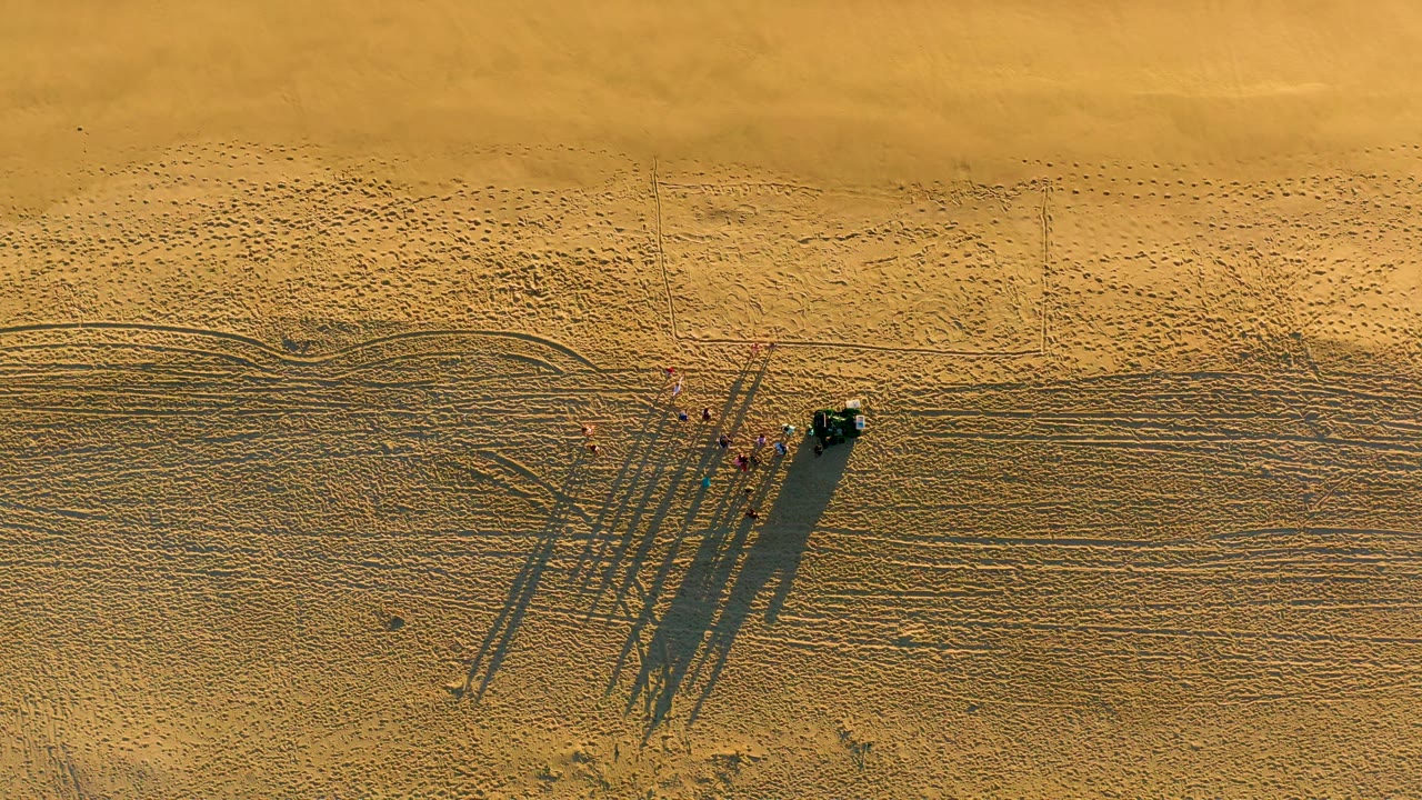 Aerial beach view with clustered people, vehicle, and shadows on patterned sand.