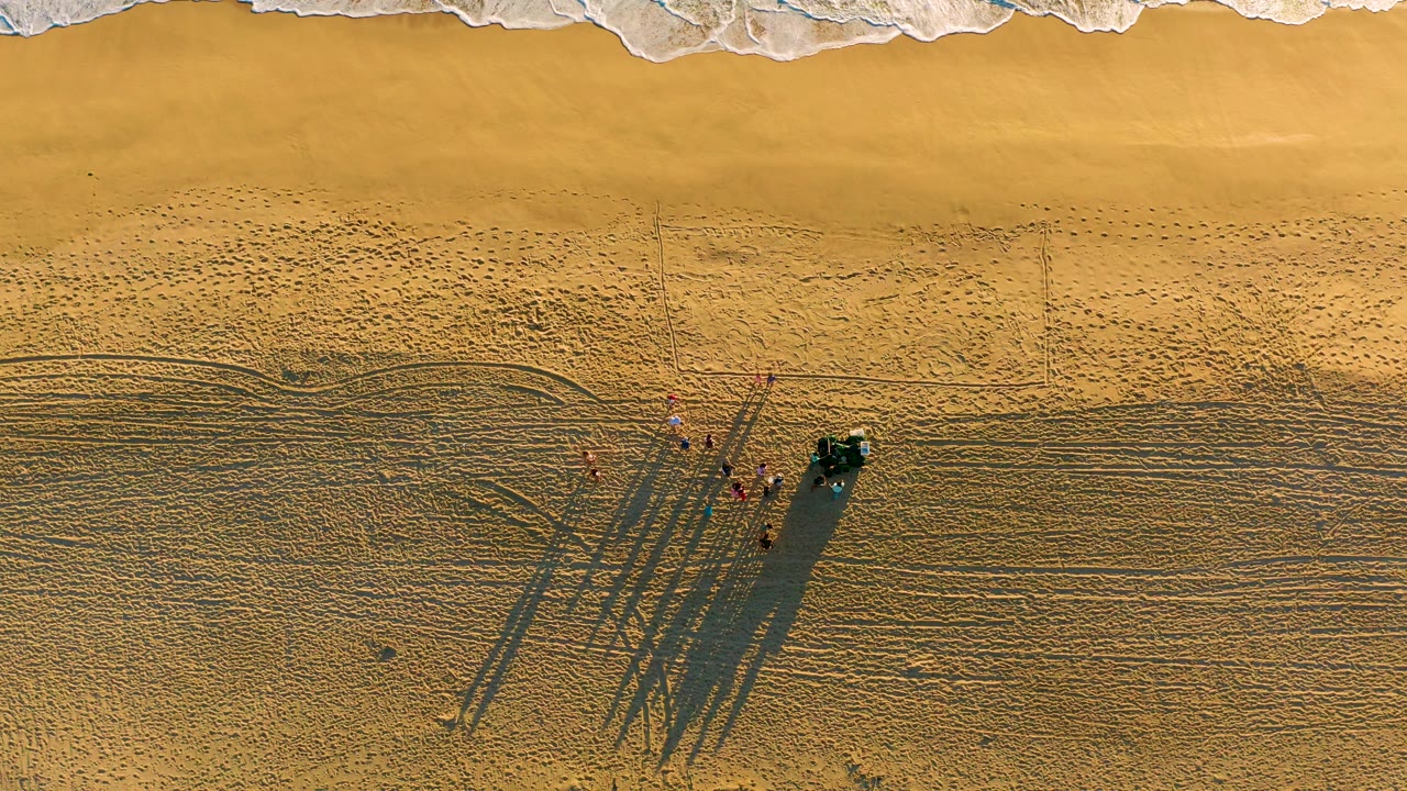 Overhead view at Diamante Cabo San Lucas showing a small group of people beside a dark vehicle on golden sand, with white surf along the top edge.