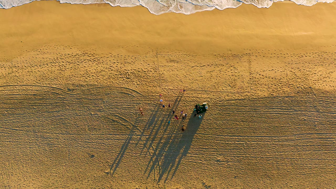 Overhead beach view with waves, people, vehicle, and long shadows.