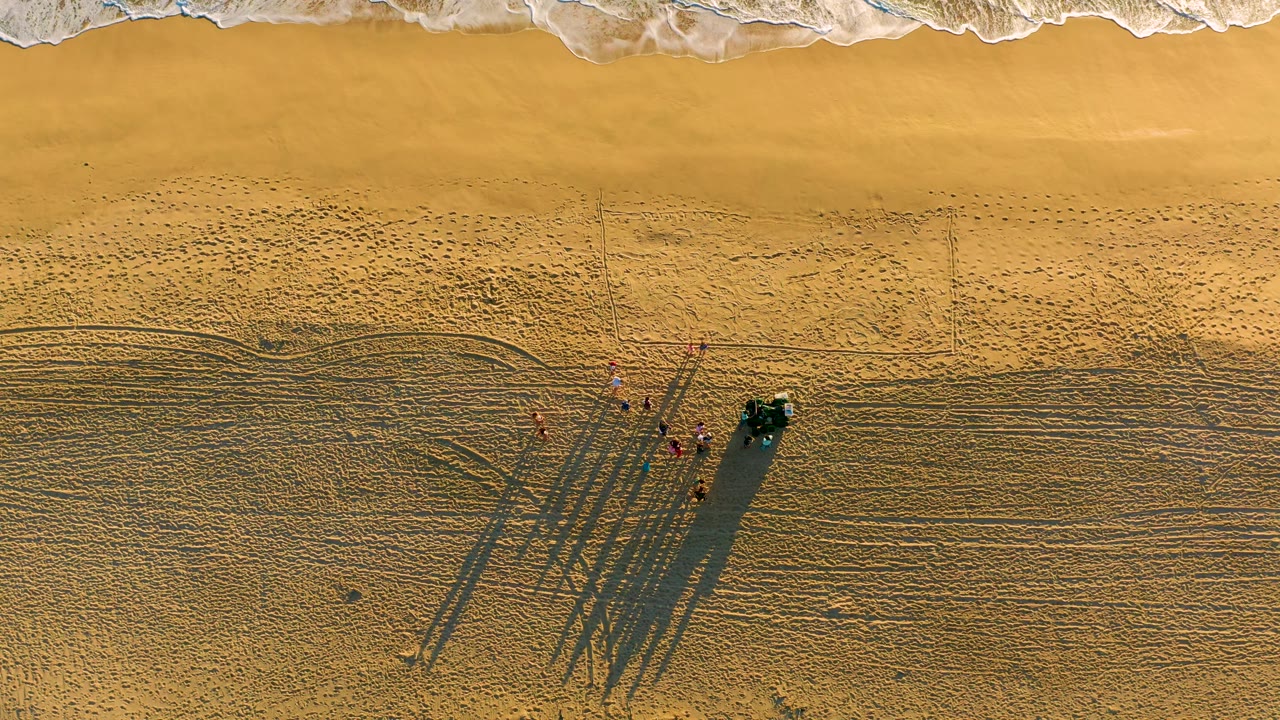 Top-down view at Diamante Cabo San Lucas with advancing white water near the upper frame and the people clustered by a dark vehicle on textured sand below.