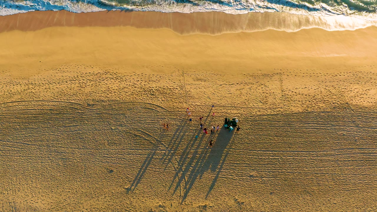At 0:02, foamy ocean water runs across the top edge while visible workers in red shirts gather beside the vehicle on textured sand at Diamante Cabo San Lucas.