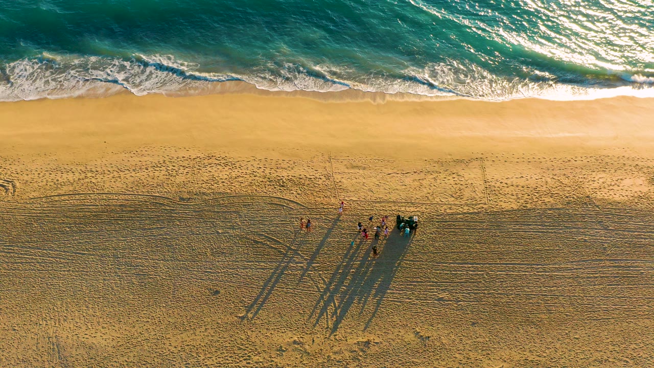 Overhead view of Diamante Cabo San Lucas beach, with turquoise water above the surf line and a small group on the sand casting long shadows beside dark beach equipment.