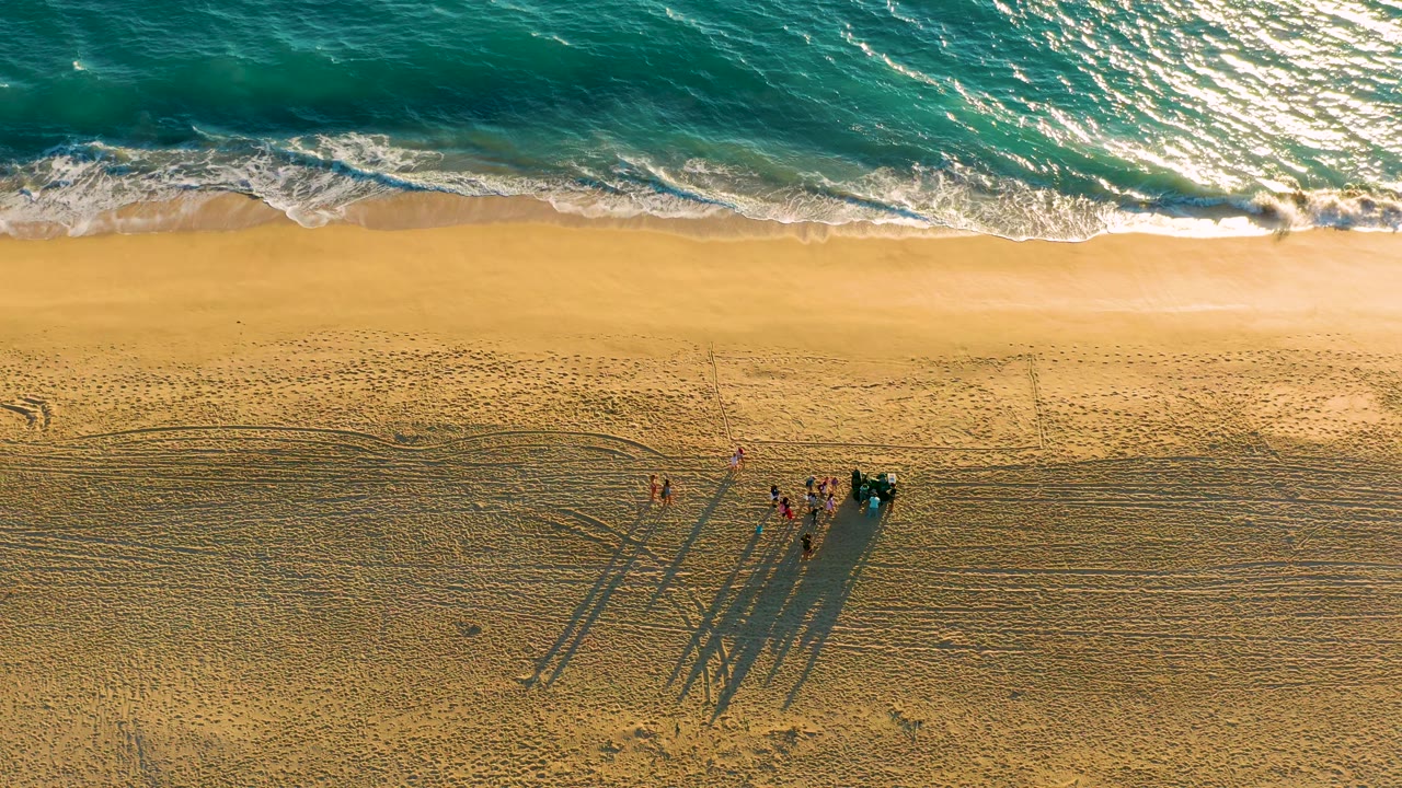 Top-down Diamante Cabo San Lucas beach view with bright white foam at the waterline and a small cluster of people near a dark vehicle or equipment on the lower sand.