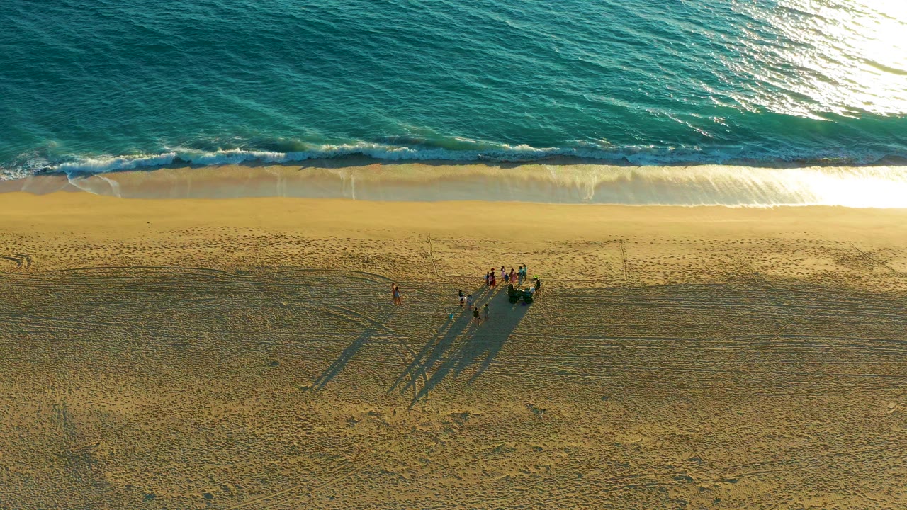 Aerial view of Diamante Cabo San Lucas beach with turquoise ocean at the top, pale surf on the shoreline, and a small group of people casting long shadows on the sand.