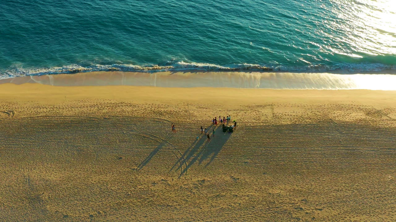 Aerial frame over Diamante Cabo San Lucas with rippled turquoise water, a thin white wave band, and several people clustered on the sand below the shoreline.