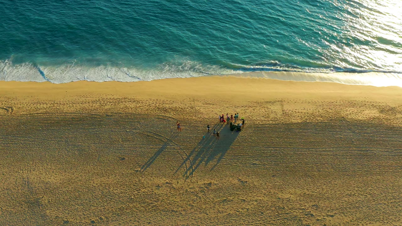 At 0:00, an overhead view at Diamante Cabo San Lucas shows turquoise surf along the top edge and a small group of people with long shadows on the lower sand.
