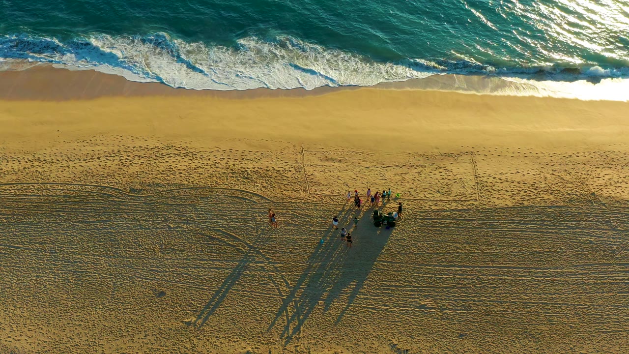 Aerial frame over Diamante Cabo San Lucas shows turquoise surf at the top, broad sand below, and a small group of people casting long shadows near the lower beach.