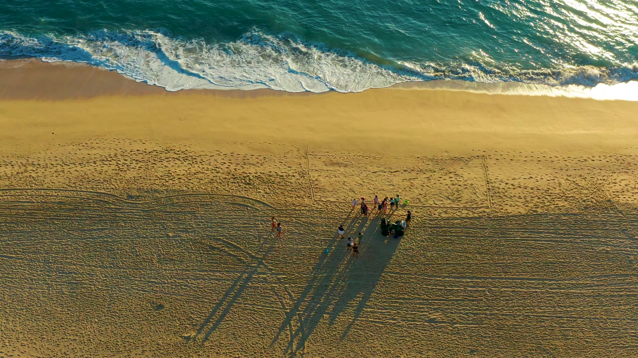 At 0:02, the aerial view holds ocean across the top and textured sand below, with the group of people and their elongated shadows shifted toward the right side of the frame.