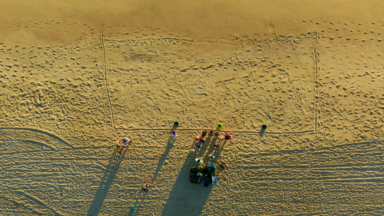 Drone overhead frame from Diamante Cabo San Lucas showing the clustered people and small vehicle lower in frame, with broad rippled sand texture and faint tire curves surrounding them.
