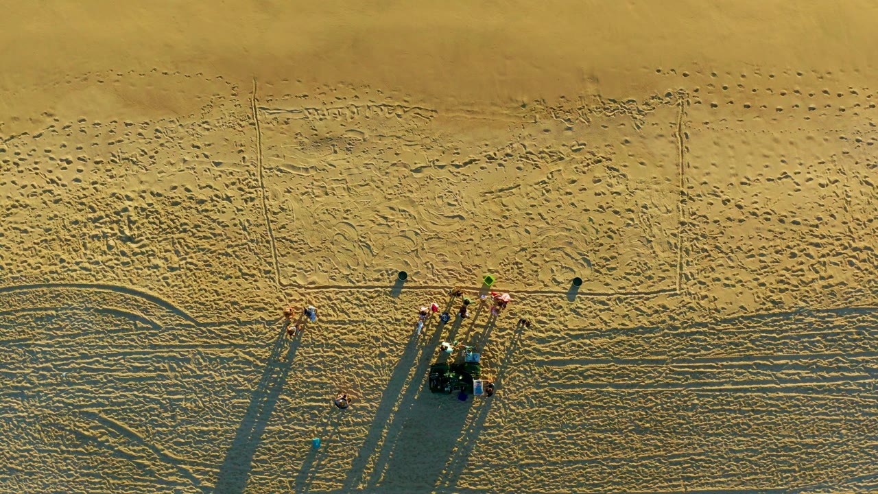 Overhead view in Diamante Cabo San Lucas showing several small people near beach maintenance equipment, tire marks, and long shadows across textured sand.