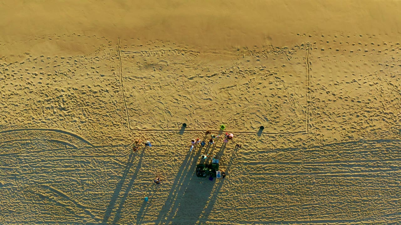 Overhead beach frame in Diamante Cabo San Lucas showing the crew still near the equipment cluster while individual shadows stretch across the lower sand.