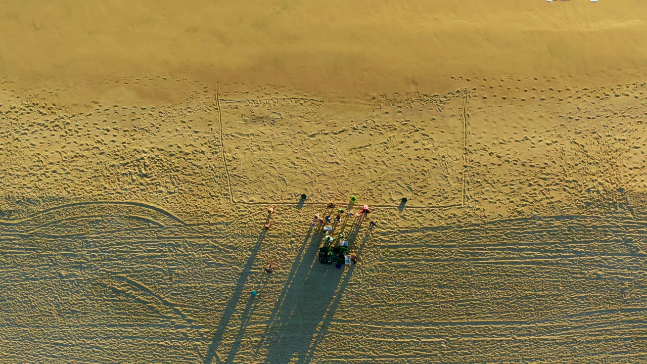 Top-down Diamante Cabo San Lucas beach view showing the group clustered at the vehicle, a few separated figures, broad rippled sand, and long shadows across the lower half.