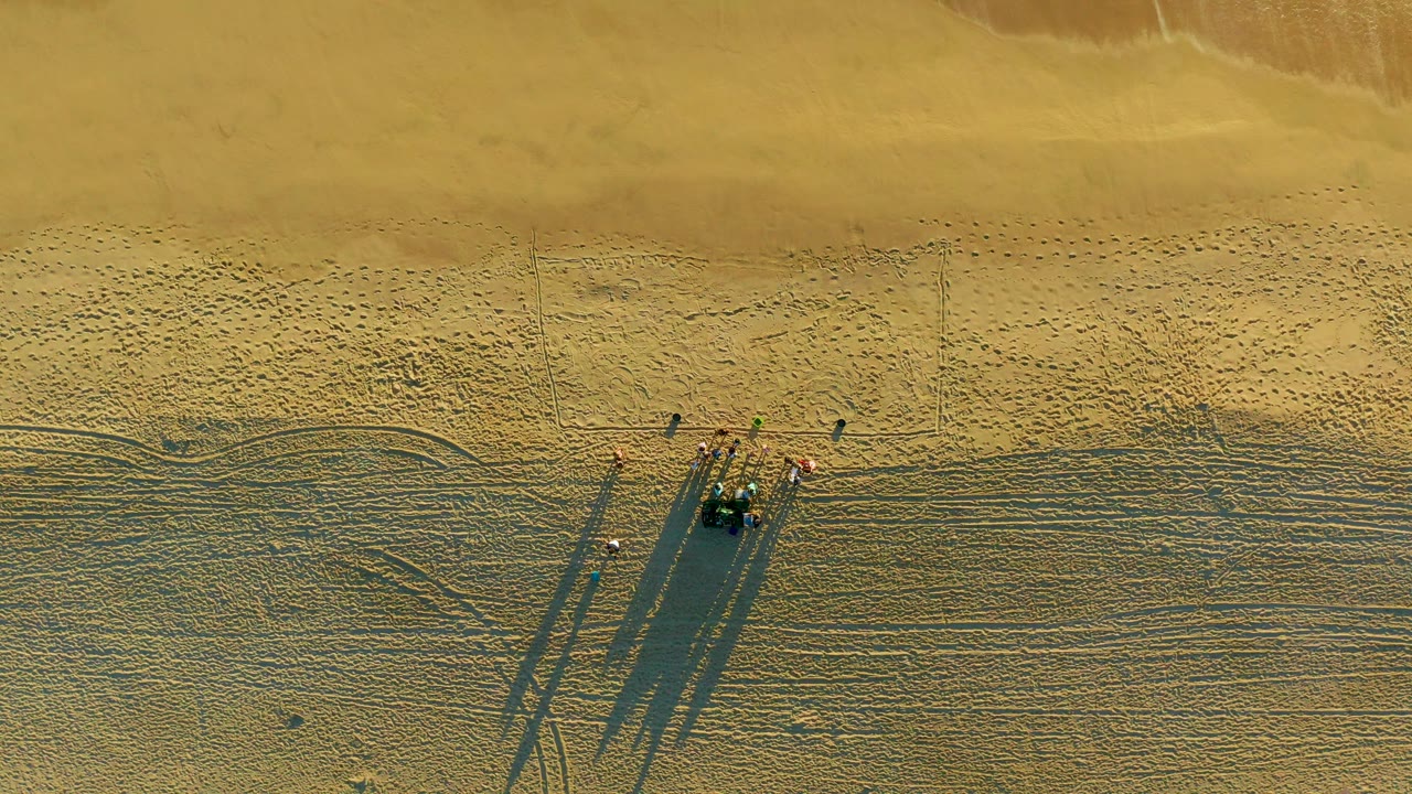 Straight-down aerial frame over Diamante Cabo San Lucas beach showing a small group on rippled sand with long shadows and faint shoreline tone near the top.