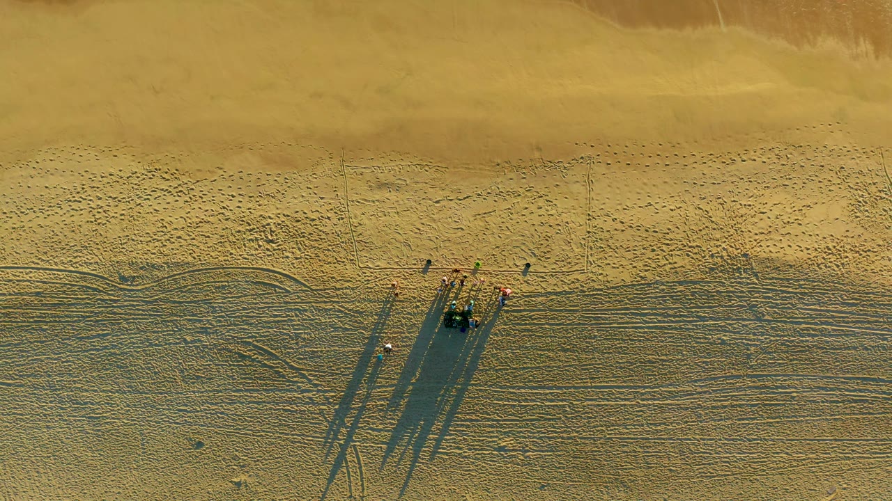 Drone frame over the Diamante Cabo San Lucas shoreline where the walking group remains on open sand and a small line of white surf enters near the upper right edge.