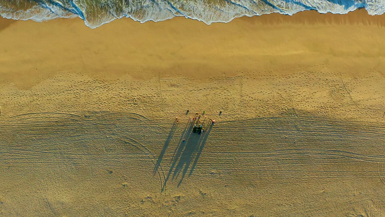Overhead view at Diamante Cabo San Lucas showing surf along the top of frame, golden sand below, and a small group of beachgoers casting long shadows near tire tracks.