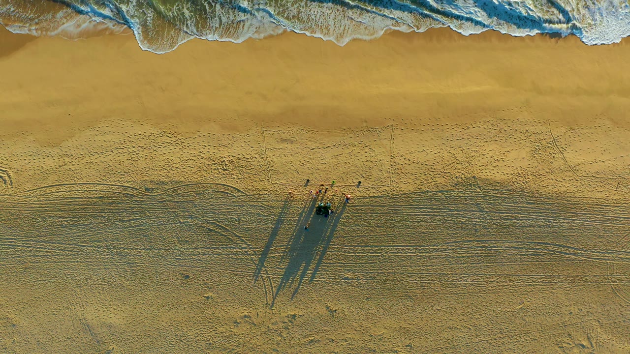 Top-down Diamante Cabo San Lucas shoreline view showing receding surf at the top, smooth wet sand beneath it, and a small group with elongated shadows lower in frame.