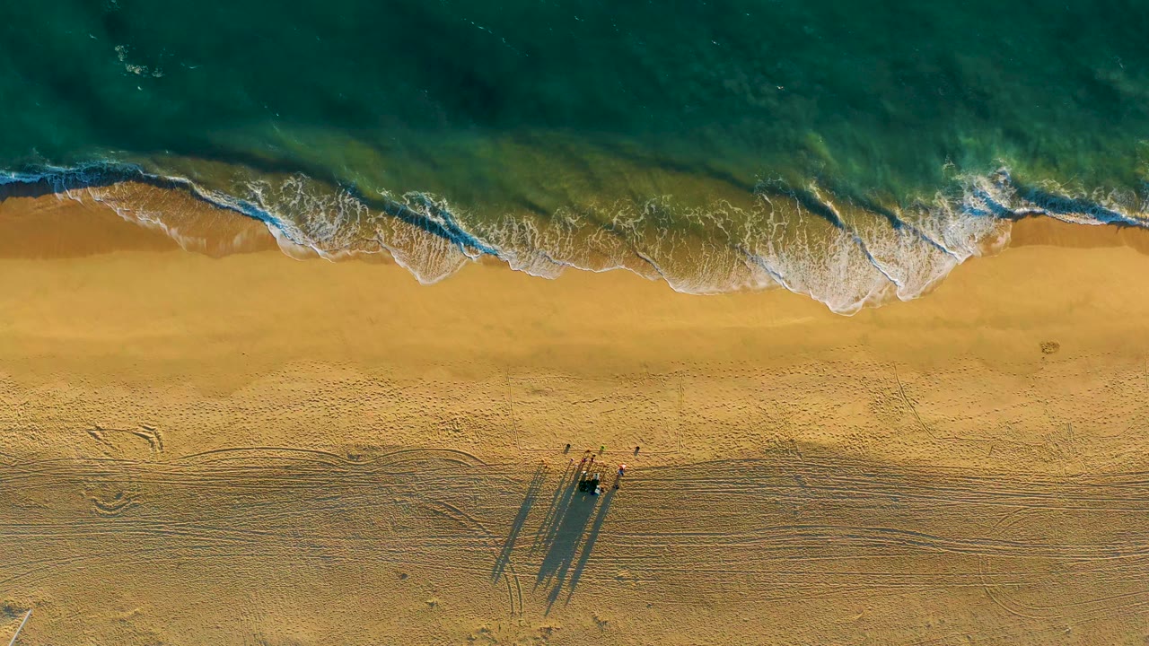 Overhead view of teal waves and white foam meeting golden sand at Diamante Cabo San Lucas, Mexico, with no visible people.