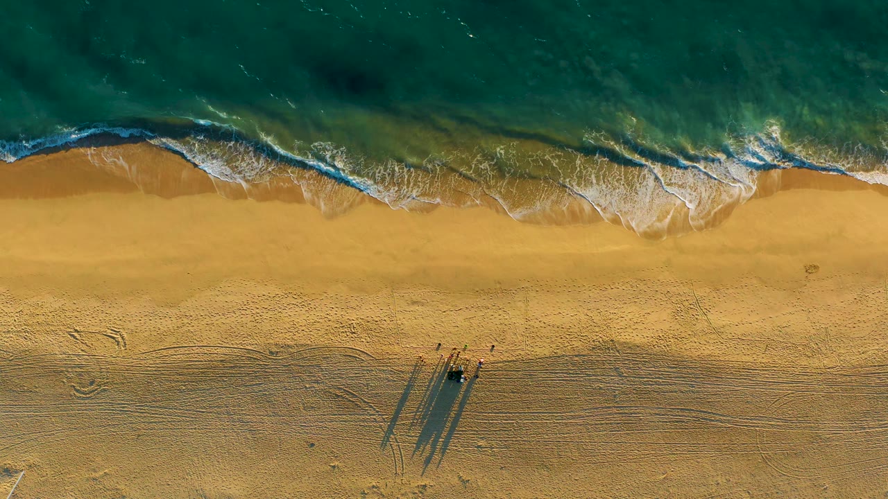 Top-down surf washes toward golden sand while a small cluster of dark beach objects casts long shadows near the lower part of the frame, with no visible people discernible.
