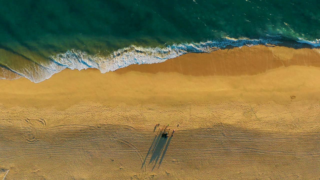 Overhead view of turquoise waves reaching golden sand at Diamante Cabo San Lucas, with white foam near the upper shoreline and no visible people.