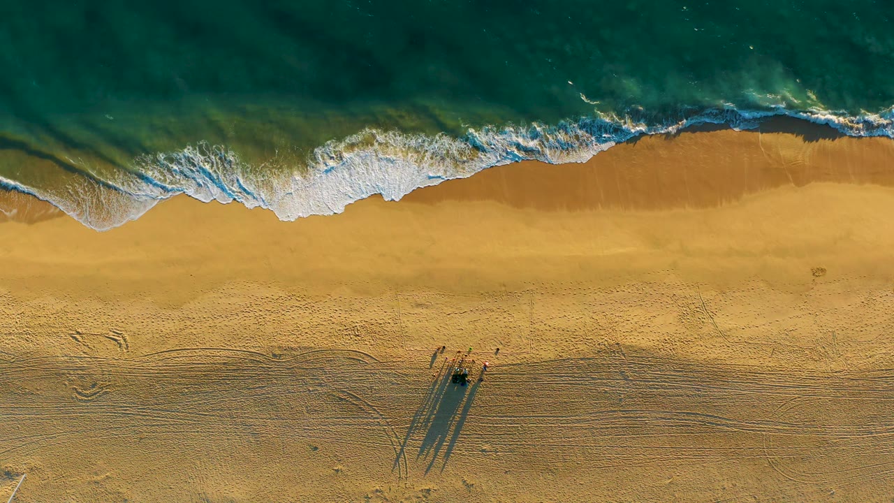 An aerial frame shows a wider sweep of foamy surf rolling onto the empty sand at Diamante Cabo San Lucas, with no visible people.
