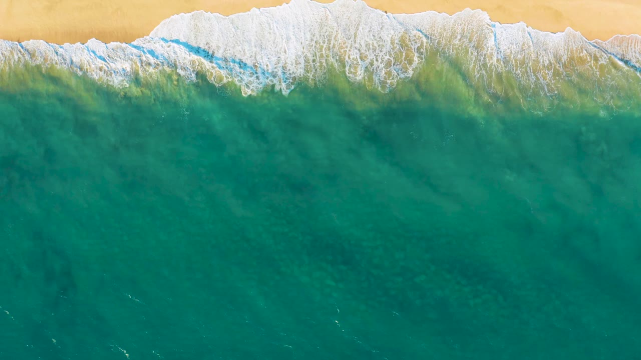 Overhead view of turquoise water and white surf meeting tan sand at Diamante Cabo San Lucas, with no visible people.