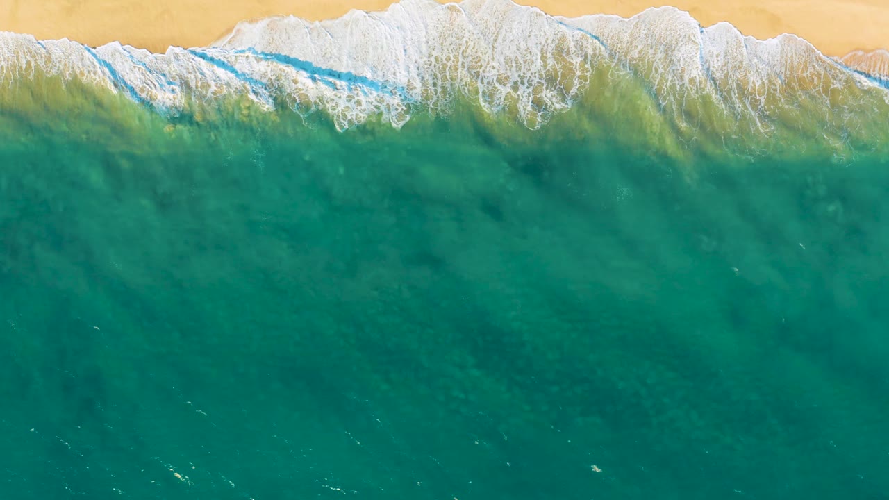 Top-down Diamante Cabo San Lucas beach view with deep turquoise water below a narrow band of breaking surf and tan sand above, with no visible people.