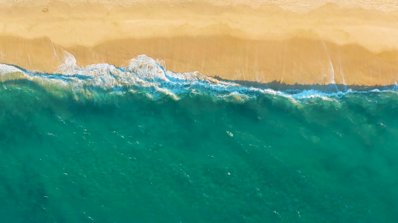 A broad band of green-blue water sits below pale sand as white surf spreads across the shoreline at Diamante Cabo San Lucas, with no visible people.