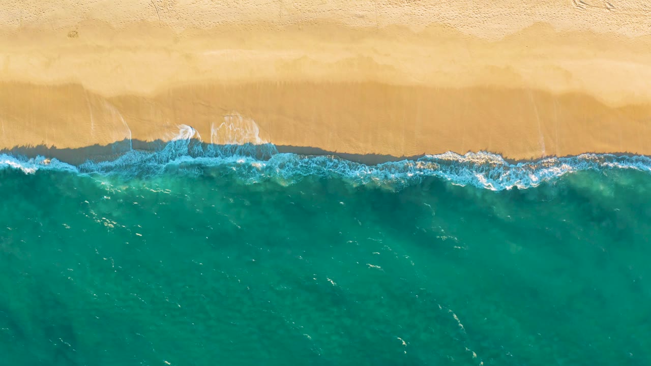 Turquoise water fills the lower frame while a thin line of white foam washes against the upper sandy beach at Diamante Cabo San Lucas, with no visible people.