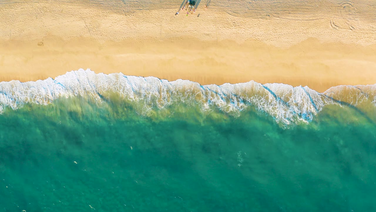 High overhead view of tan sand and turquoise surf at Diamante Cabo San Lucas, with white foam along the shoreline and no visible people.