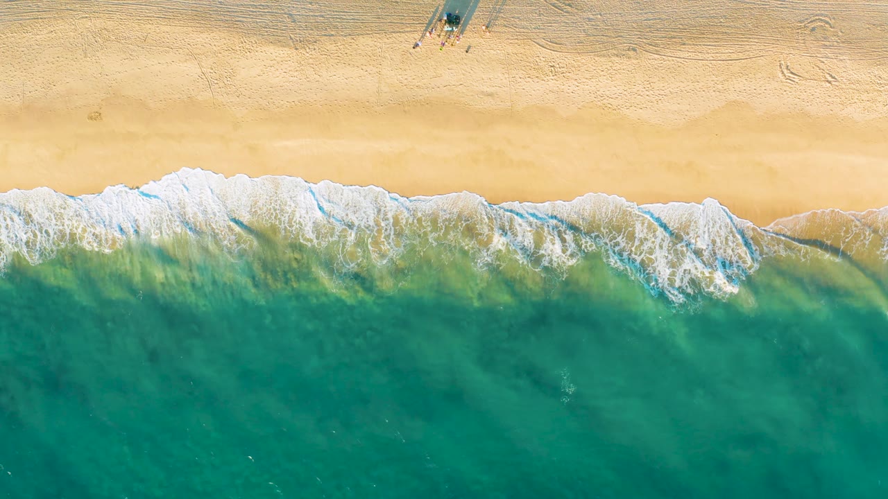 Top-down view of Diamante Cabo San Lucas surf with a broad white foam line between pale sand and green-blue ocean water, with no visible people.
