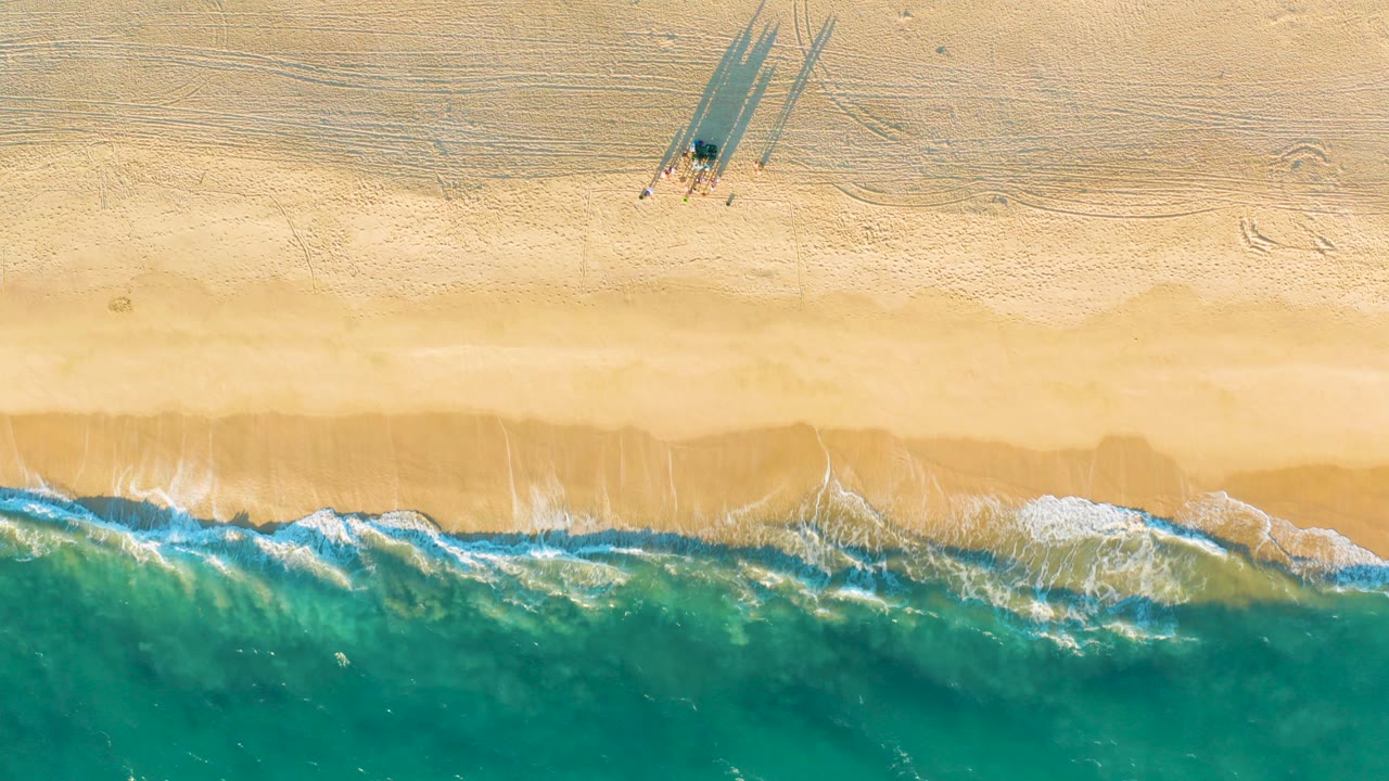 Straight-down aerial frame of Diamante Cabo San Lucas beach with tan sand above, turquoise waves below, white foam at the shoreline, and a tiny shadowed figure or object near the upper sand.